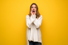 Blonde woman over yellow wall shouting and announcing something