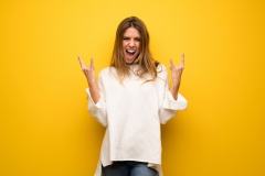 Blonde woman over yellow wall making rock gesture