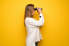 Blonde woman over yellow wall and looking in the distance with binoculars