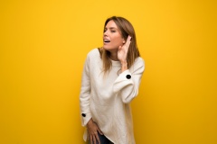 Blonde woman over yellow wall listening to something by putting hand on the ear