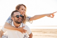 Photo of unshaven cheerful man smiling and giving piggyback ride seductive woman while walking on sunny beach
