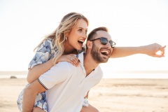 Photo of handsome happy man smiling giving and piggyback ride seductive woman while walking on sunny beach