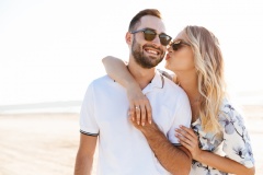 Photo of blonde young woman kissing and hugging handsome man while walking on sunny beach