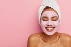 Close up portrait of young female model applies homemade facial clay mask, has white towel wrapped around head, keeps eyes shut, smiles happily, models against pink background. Beauty treatment
