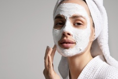 Young woman in a towel applying facial white mask at her face at home, isolated on a gray background. Skin care concept. Advertising poster of natural cosmetics