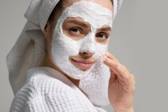 Pretty woman with towel on head apply face mask for acne doing morning beauty routine isolated on studio gray background. Healthy young skin care concept