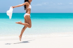 Beach ready bikini body - sexy slim legs and toned thighs and butt. Suntan happy woman jumping in freedom on white sand with sun hat. Weight loss success or epilation concept.