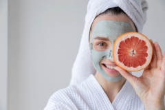 Young woman getting mud clay mask on face and holding grapefruit slice.