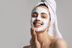 Smiling pretty girl in towel applying eco-friendly skincare mask for acne on face isolated on gray background. Morning beauty routine. Skincare treatment concept