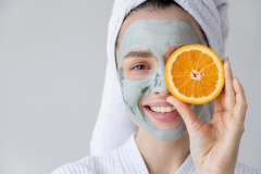 Smiling happy woman with grey clay facial mask applied on face holding orange citrus fruit slice.