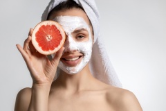 Beautiful young smiling woman with white clay mask  holding grapefruit near face.