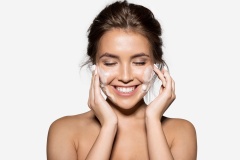 Portrait of cheerful laughing woman applying foam for washing on her face. Lovely brunette with attractive appearance. Skincare spa relax concept. Isolated on grey background
