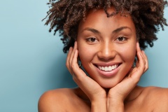 Close up portrait of relaxed black woman has gentle skin after taking shower, satisfied with new lotion, has no makeup, smiles tenderly, shows perfect teeth, stands shirtless against blue background