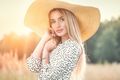 Beautiful model girl posing on a field, enjoying nature outdoors in wide brimmed straw hat. Beauty blonde young woman with long straight blond hair closeup portrait