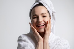 Happy laughing young woman wearing bathrobe and head towel with nude makeup touching face.